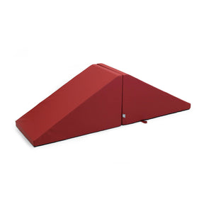 Introducing the Mega Wedge Play Triangles by Foamnasium: a vibrant red foam wedge featuring a fold in the middle that forms a triangular incline. This durable foam playset is wrapped in smooth, hard-wearing fabric for longevity. A small handle on one side ensures easy folding and transportation. The product is showcased against a white background.
